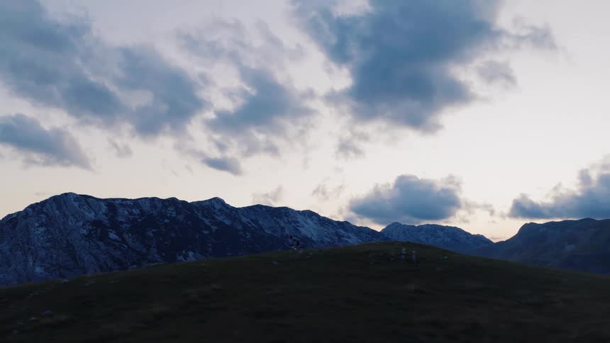 An epic aerial pan reveals a mountain biker silhouetted against a vast mountain range and cloudy sky at dusk