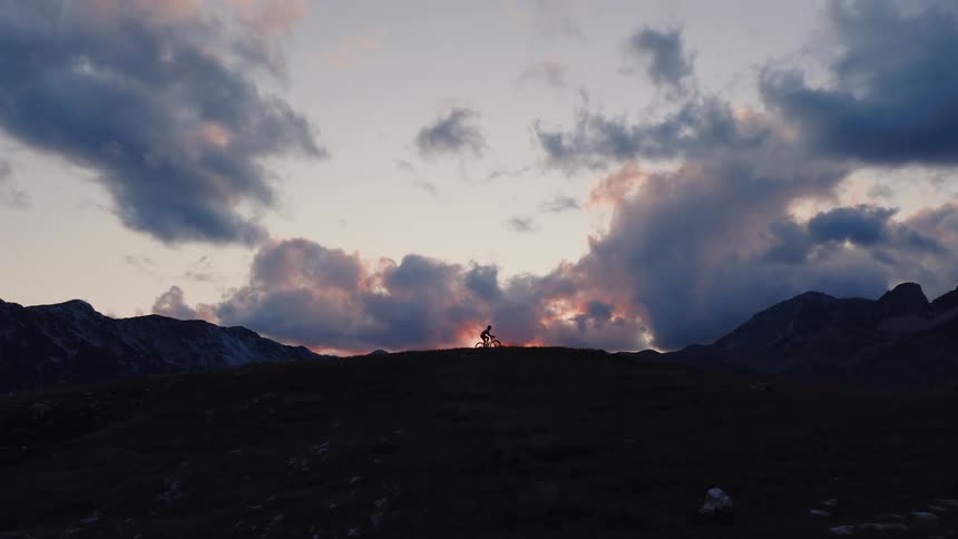A lone mountain biker rides along a mountain ridge in slow motion, silhouetted against a dramatic sunset sky