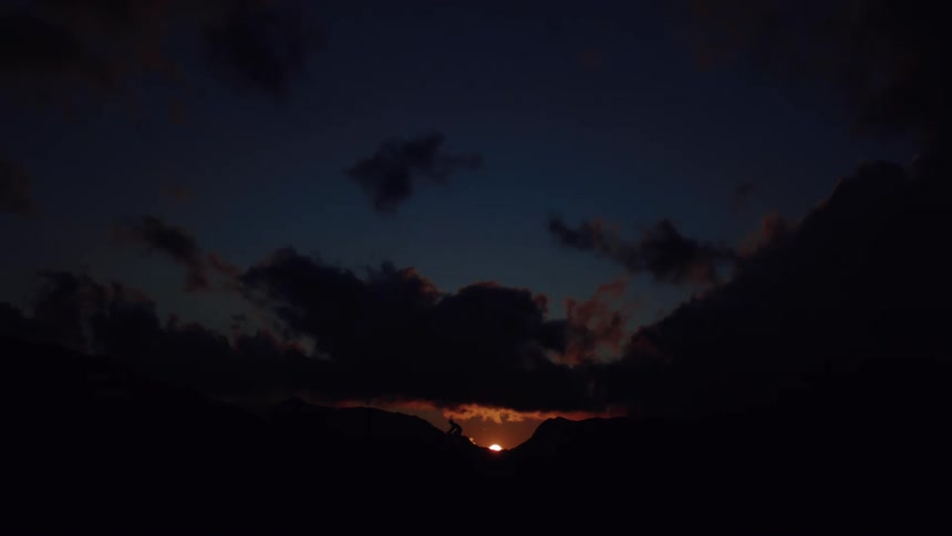 A sliver of the setting sun is visible between silhouetted mountains against a dark, cloudy sky