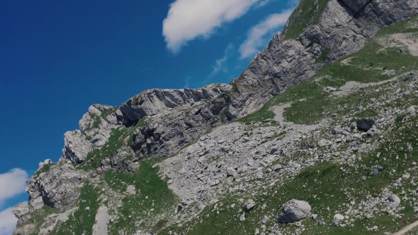 An aerial drone shot pans up a steep, rocky mountain slope with patches of green grass under a clear blue sky
