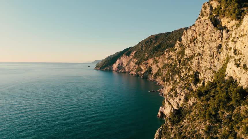 Slow aerial flight along the rugged Italian coastline as the warm morning sun illuminates the dramatic cliffs and calm sea