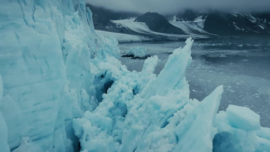 A dramatic aerial shot pulls back along a massive blue glacier wall to reveal the vast, icy landscape of an Arctic fjord and distant mountains