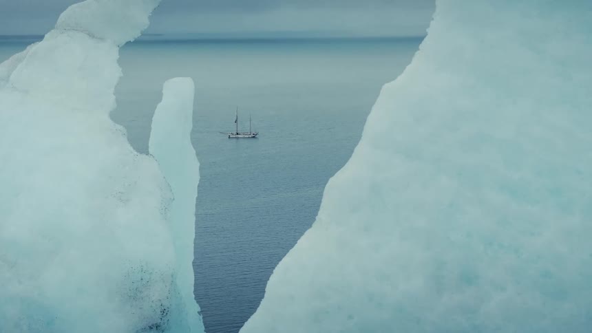 A sailboat is seen cruising through the arctic sea, framed by massive blue icebergs