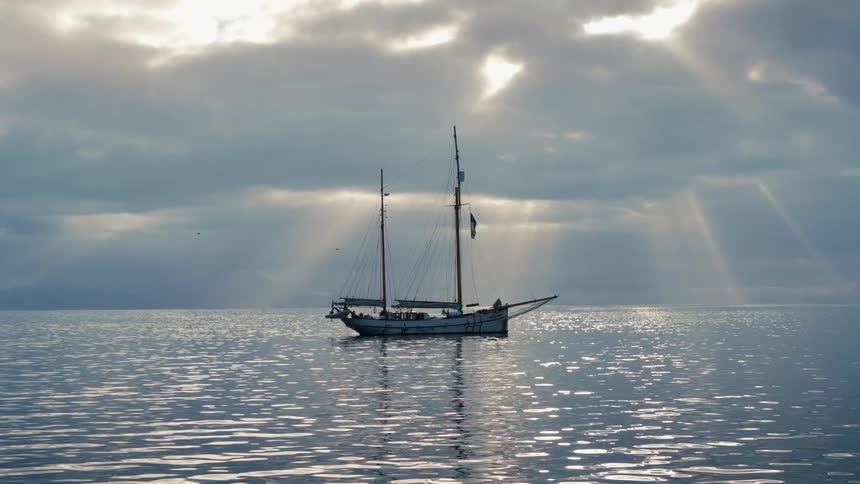 A classic two-masted schooner is silhouetted on a tranquil sea as sun rays pierce through the overcast sky