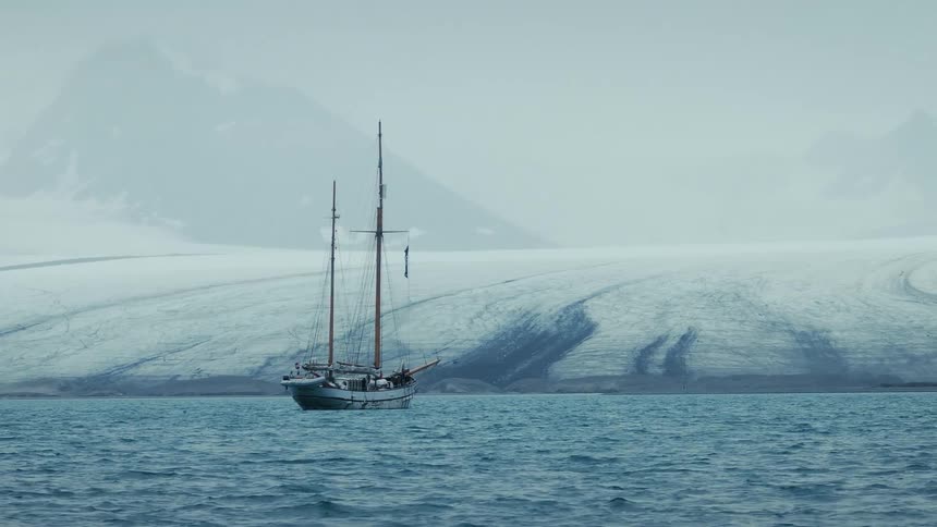 A classic sailboat floats peacefully on the choppy arctic sea with a massive glacier in the background
