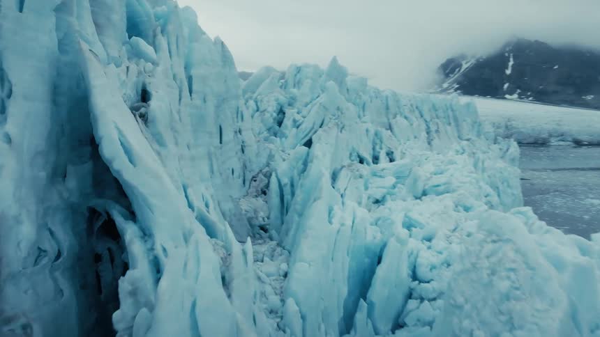 A slow, dramatic aerial shot tracks along the immense, jagged wall of an arctic glacier meeting the icy ocean