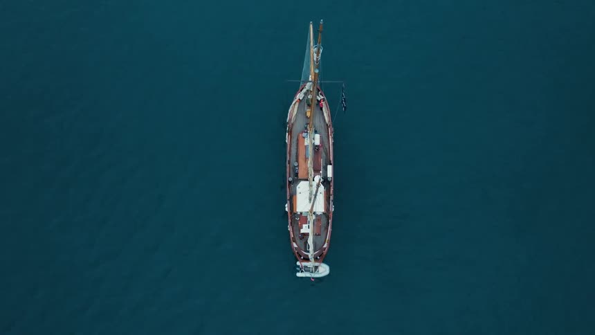 A classic wooden pirate ship with a Jolly Roger flag floats peacefully in the deep blue ocean, captured from a direct top-down aerial view