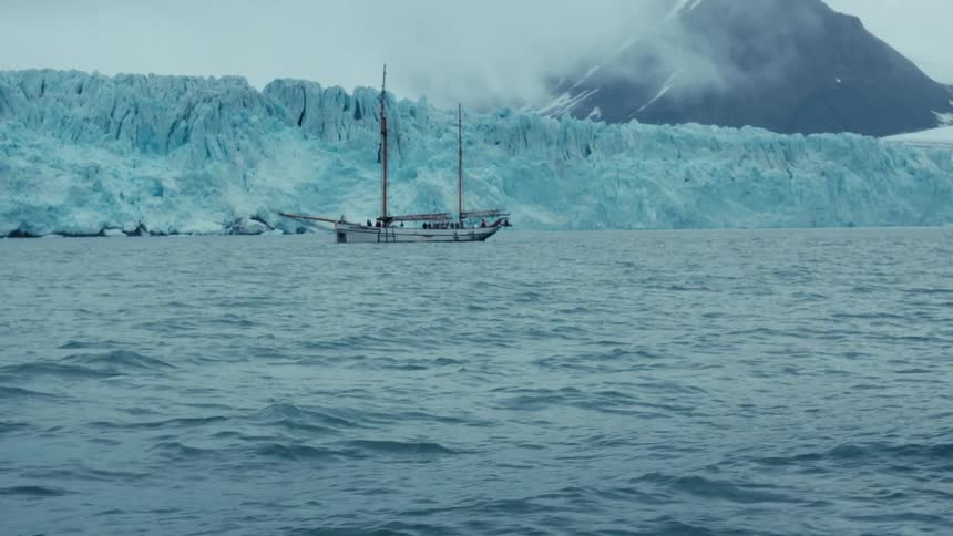 A slow tilt-up reveals a majestic sailboat floating in the arctic ocean in front of a giant glacier wall