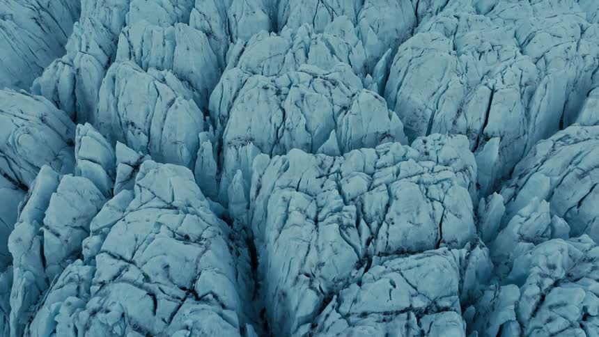 An aerial view slowly zooms in on the jagged, cracked surface of a vast blue glacier