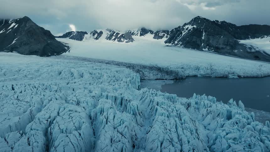 A stunning wide shot reveals a massive, crevassed glacier flowing into a fjord at the base of snow-covered mountains under a dramatic sky