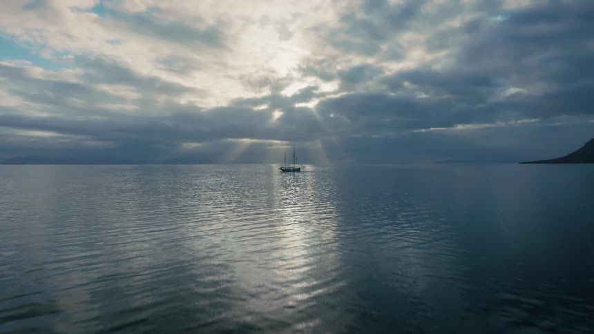 A lone sailboat floats peacefully on a calm ocean as beautiful sunbeams break through the dramatic, cloudy sky