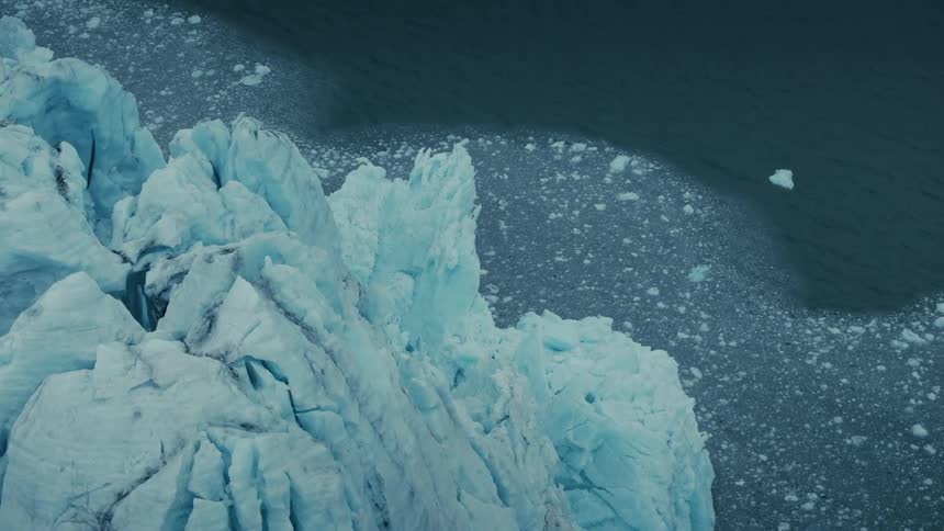 An overhead aerial view captures the slow, powerful calving of a massive blue glacier into the dark ocean water