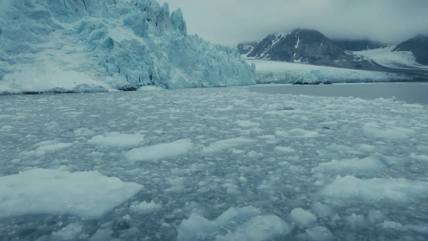 A slow tilt up from icy waters reveals a majestic glacier and snowy mountains in a vast arctic landscape