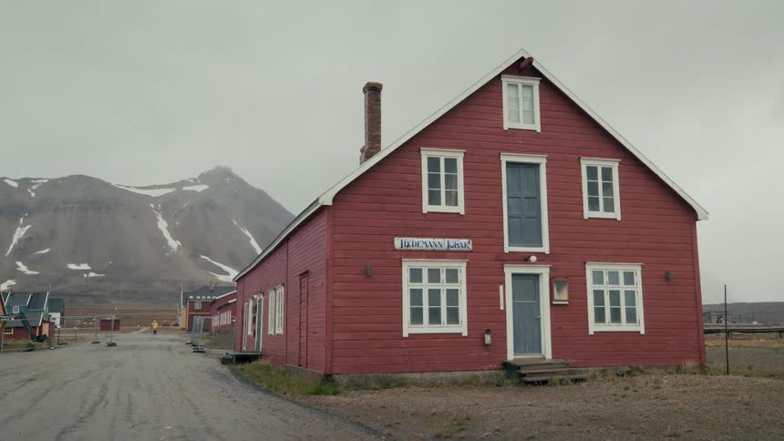 A static shot of a classic red wooden house in a remote northern village, with snow-capped mountains in the background under an overcast sky