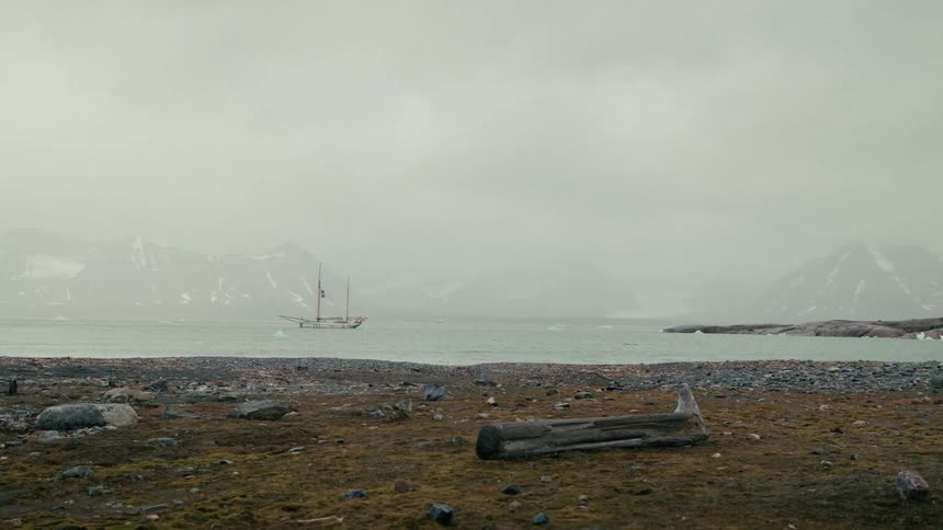 A vintage sailing vessel floats in a foggy arctic fjord, seen from a desolate rocky shoreline with snowy mountains in the background