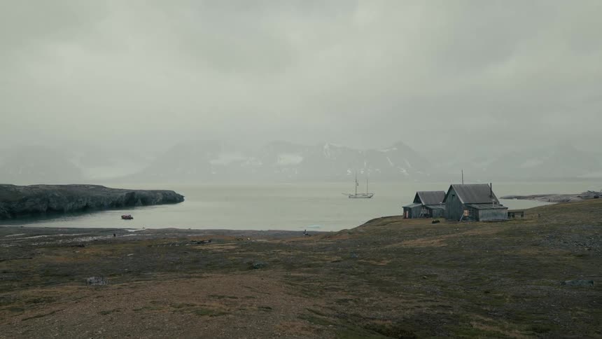 Weathered wooden cabins stand on a desolate northern shoreline as a classic sailing ship rests in the foggy bay