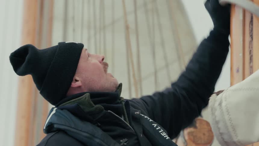 A crew member wearing a beanie and life vest carefully raises the sail on a wooden mast in slow motion