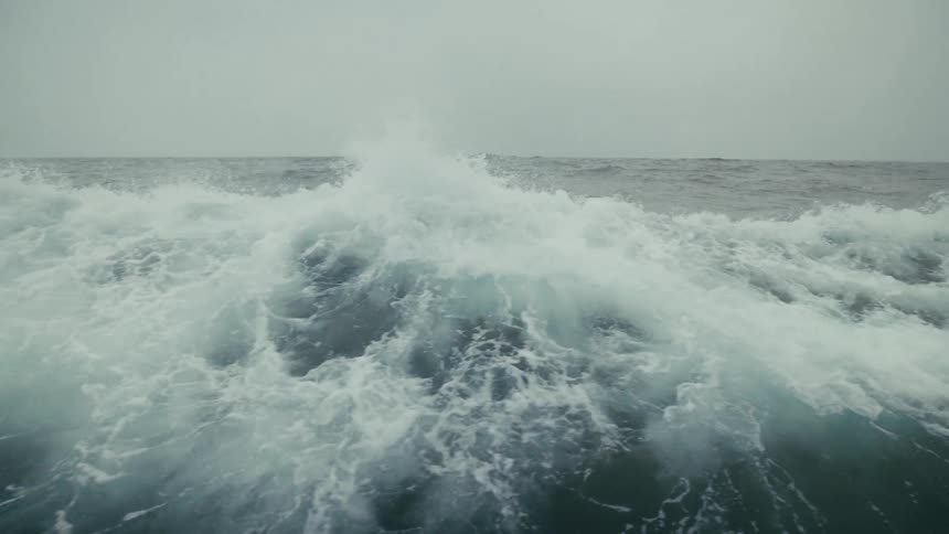 A dramatic close-up of a powerful, frothy ocean wave swelling and cresting in slow motion under a gray sky