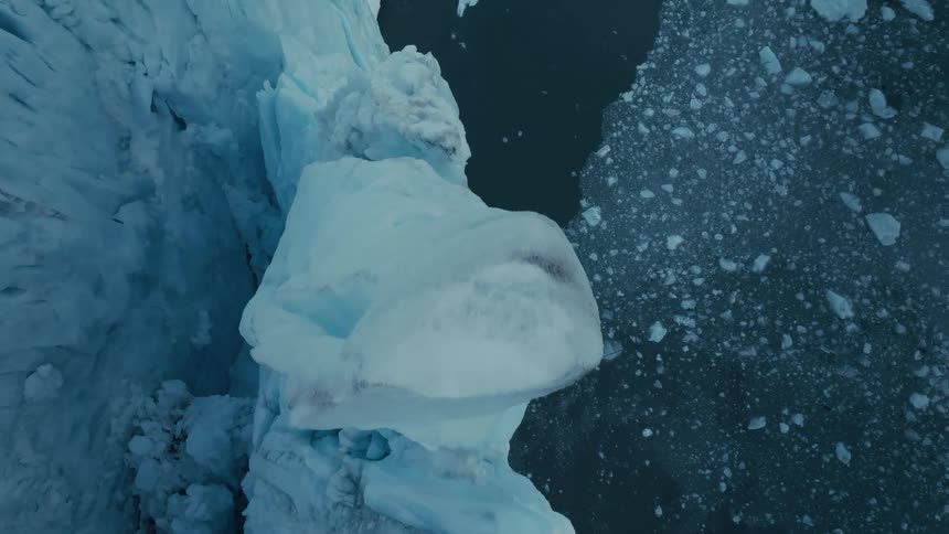 A dramatic top-down aerial view captures the slow, powerful process of a massive glacier calving into the frigid arctic water below