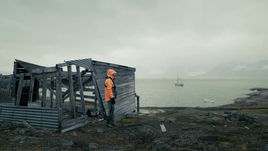 A lone explorer in an orange raincoat walks out of a dilapidated wooden shack on a desolate coastline with a sailboat on the water in the distance