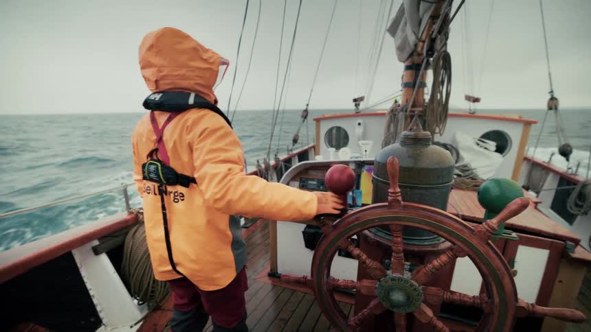 A sailor in an orange rain jacket takes the helm and steers a classic wooden sailboat across the choppy open ocean