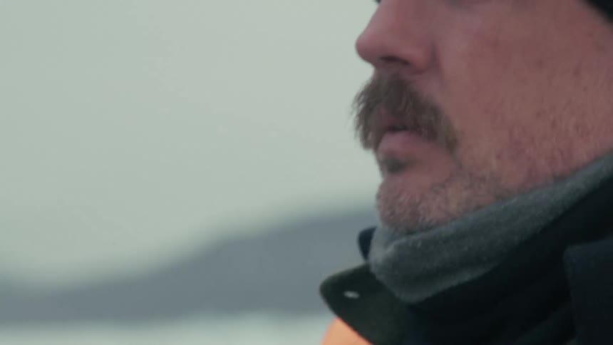 A man with a mustache wearing a black beanie and an orange vest speaks into a radio on an overcast day