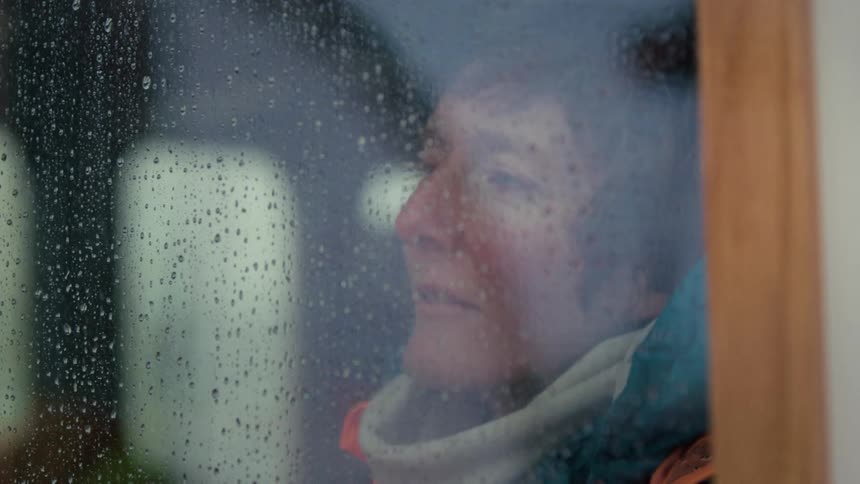 A contemplative woman watches rain fall from inside, her face reflected in the wet window pane