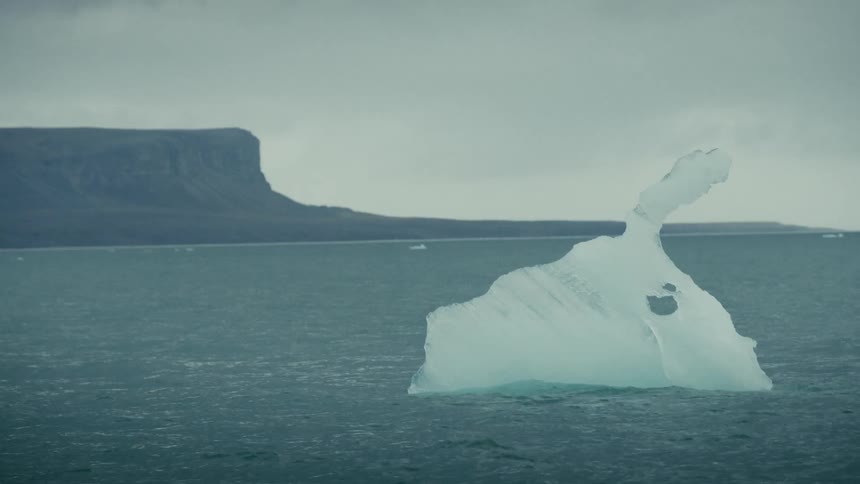 A lonely iceberg drifts in the grey, choppy sea under an overcast sky, with a mountainous coastline in the distance
