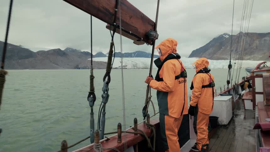 On the wet deck of a traditional sailboat, two explorers in rain gear watch as they approach a vast glacier in the Arctic