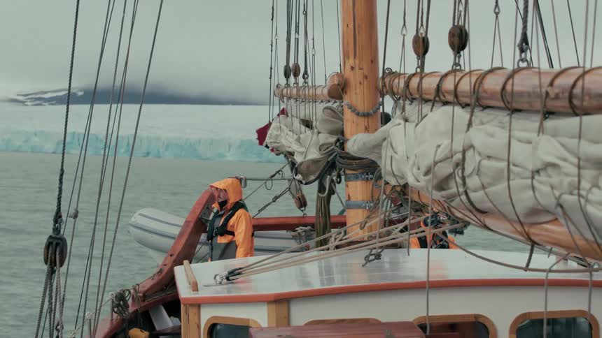 A sailor in a bright orange jacket stands on a wooden sailboat as it navigates past a massive arctic glacier on a cold, overcast day