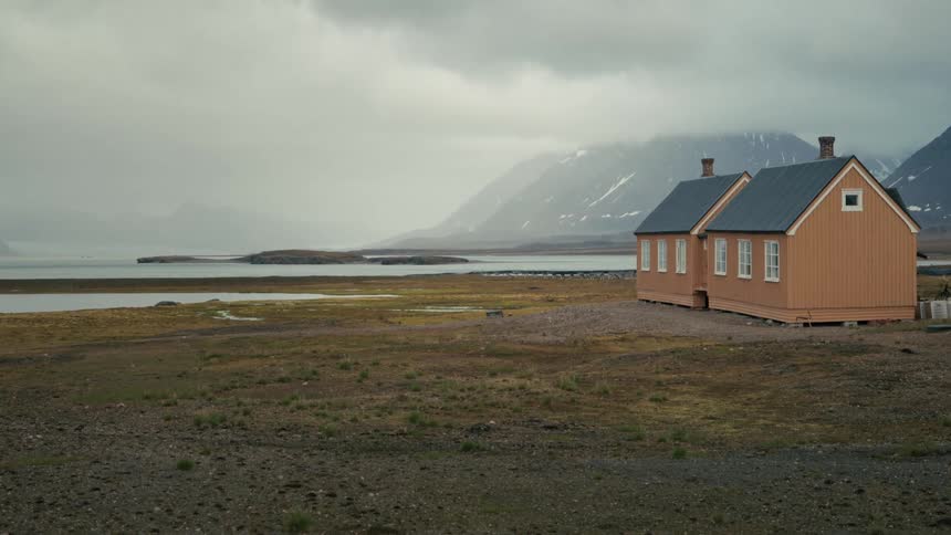 Two isolated yellow houses sit on a desolate shore with misty, snow-capped mountains in the background