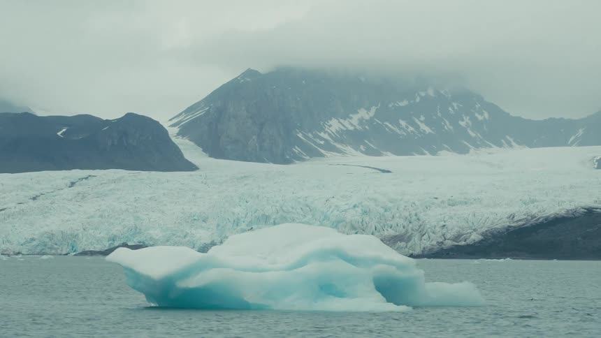 A stunning view of a bright blue iceberg floating in arctic waters with a massive glacier and misty mountains in the background