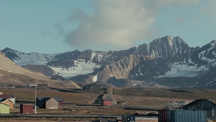 A small, colorful arctic village sits in a vast, barren valley beneath majestic, snow-covered mountains