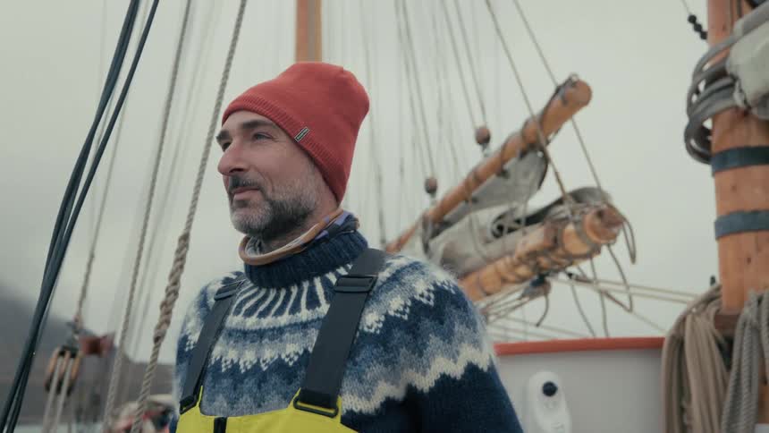 A handsome sailor with a beard, wearing a red beanie and wool sweater, smiles while standing on the deck of a classic wooden sailboat