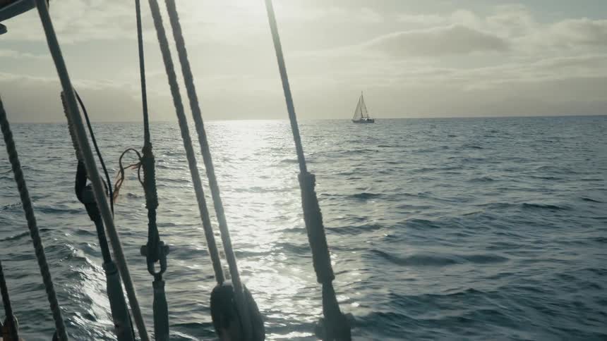 A tranquil view past the rigging of a sailboat shows a distant yacht on the glittering ocean during a hazy sunset