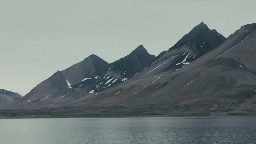 Vast, barren mountains with patches of snow stand tall over a tranquil lake under an overcast sky