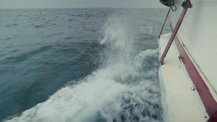 A boat's hull powerfully slices through turbulent ocean waves in dramatic slow motion on a gloomy day