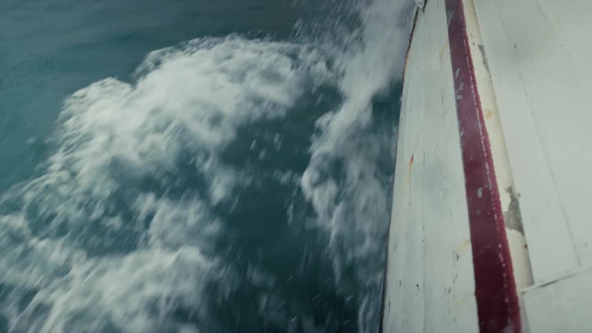 The hull of a boat slices through the turbulent ocean, creating white foamy waves on a grey day