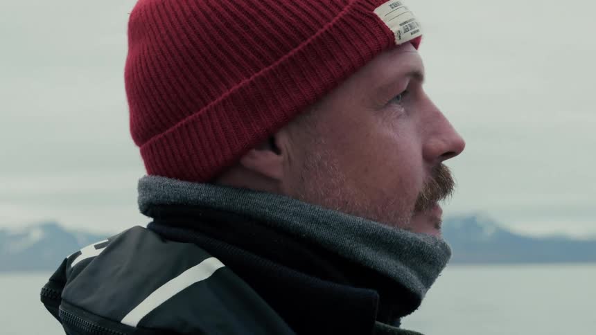 A close-up profile shot of a contemplative man in a red beanie looking out at a vast, chilly seascape with mountains in the background