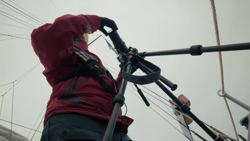 A photographer wearing a red jacket and life vest sets up a camera on a tripod on the deck of a sailing vessel