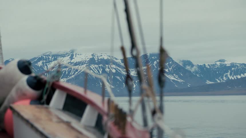 A serene, static shot of snow-capped mountains across calm water, viewed from a boat on an overcast day