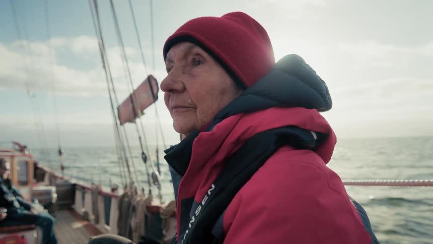 A contemplative elderly woman in a red beanie gazes out at the sea from the deck of a sailboat