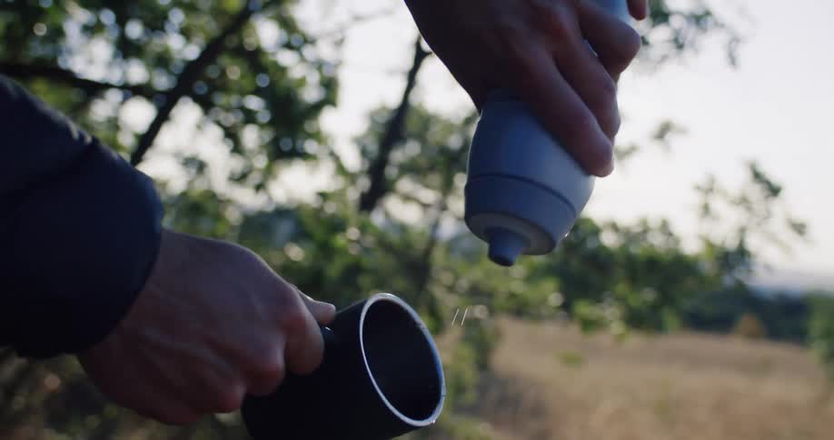 A person pours water from a bottle into a mug and then spills the rest onto the ground in a sunlit field in slow motion