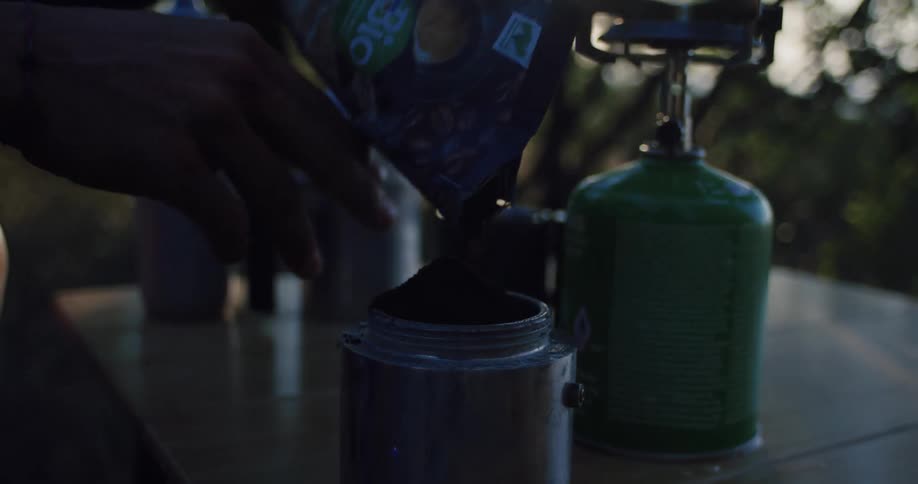A camper pours ground coffee into a moka pot in slow motion with a camping stove in the background