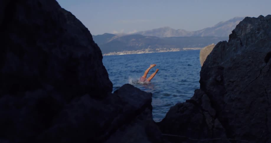 An athletic man takes a thrilling dive in slow motion from a coastal rock into the sea with mountains in the background