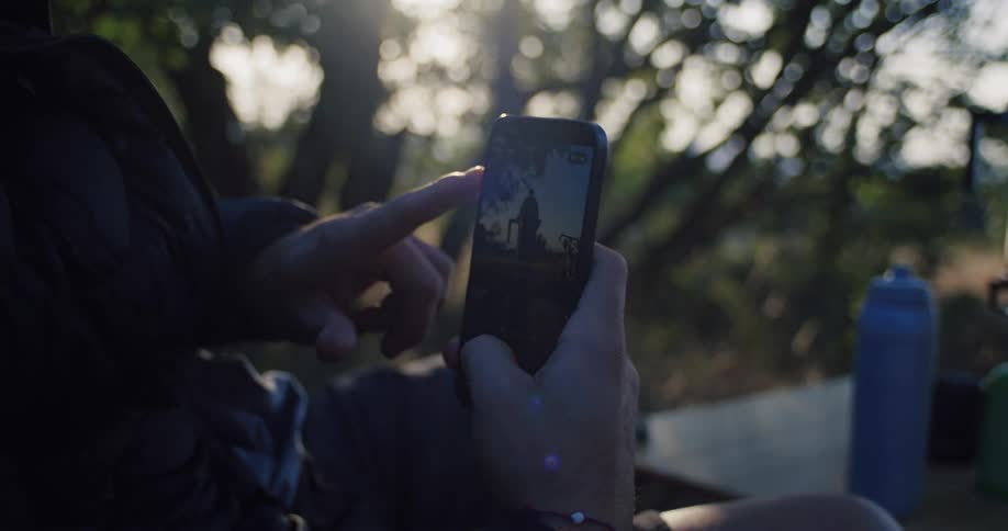 A person's hands hold a smartphone, scrolling through a video of a cyclist while resting outdoors at sunset