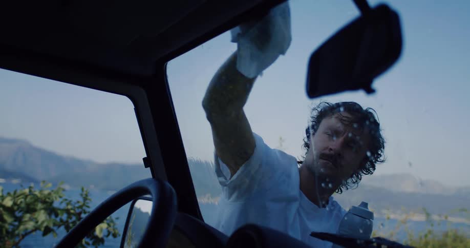 Interior slow-motion view of a man pouring water from a bottle and wiping the windshield of his car clean by a scenic coastline