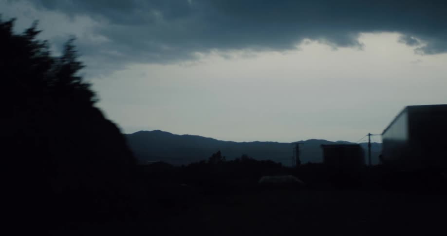 A dark, moody tracking shot captures a silhouetted industrial landscape with distant mountains under an overcast sky