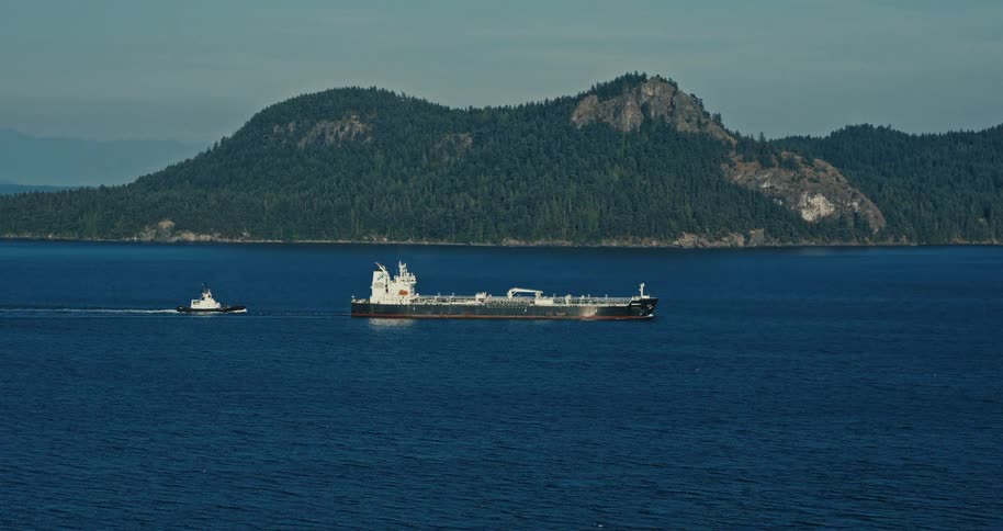A large cargo tanker ship is escorted by a tugboat across a vast blue ocean with a lush green island in the background