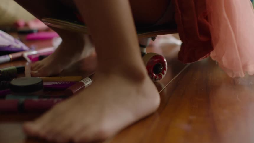 Close-up on a young child's bare feet as they playfully scoot on a skateboard across a messy wooden floor covered in makeup